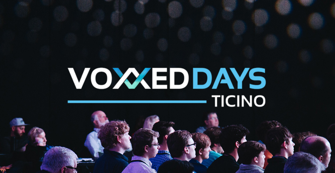 Group of attendees listening to a talk with the logo of VoxxedDays Ticino in the middle of the picture.